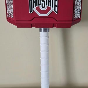 May include: A red hammer prop featuring the Ohio State logo in white, black, and red on the front face. The hammer head has silver-colored Celtic-style knotwork on the sides, and the handle is white with a red pommel at the base.