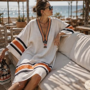 May include: A cream-colored kaftan dress featuring a deep V-neckline with orange and black striped trim. The sleeves and hem are accented with horizontal orange and black bands. A small logo in the bottom right corner reads AMARA DUNE.