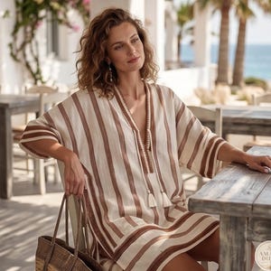 May include: A beige and brown striped tunic featuring a deep V-neckline with decorative stitching, short wide sleeves, and two hanging tassels at the center. The garment has a loose, relaxed fit with vertical stripes and a woven texture.