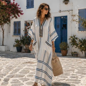 May include: A floor-length white kaftan dress featuring a V-neckline with gold and blue embroidery, wide sleeves, and two large front pockets. The garment is decorated with vertical and horizontal blue stripes and is paired with a woven straw tote bag.