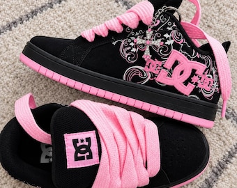 Puede incluir: Un par de zapatillas DC negras y rosas con cordones rosas gruesos, suela negra y un patrón gráfico floral blanco y rosa en el lateral. Un dije de cruz plateado con pedrería transparente está sujeto a los cordones de la zapatilla superior.