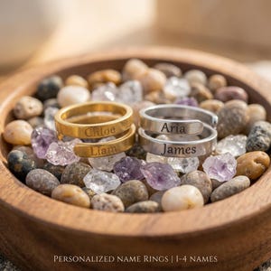 May include: Two wrap-style rings, one gold-toned and one silver-toned, each engraved with two names. The gold ring features the names Chloe and Liam, while the silver ring displays Aria and James. The text Personalized Name Rings | 1-4 Names is visible.