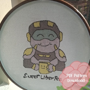 May include: A cross-stitch project in a circular wooden hoop featuring a cartoon character in purple and yellow armor holding a yellow mug with a smiley face. The text "Sweet Liber-tea" is stitched below the character. A pink flower graphic overlays the bottom right corner with the text "PDF Pattern Download".