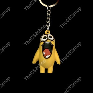 May include: A yellow keychain featuring a cartoon character with wide, surprised eyes and a large, open mouth showing a red tongue. The character is attached to a silver-colored metal chain and a circular split ring for keys.