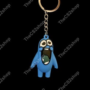 May include: A blue silicone keychain featuring a cartoon character with wide, surprised eyes and a large, open mouth showing a dark interior. The figure is attached to a silver-colored metal split ring by a short chain. The text TheCS2shop is visible.