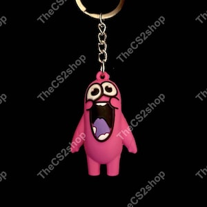 May include: A pink keychain featuring a cartoon character with a wide, open mouth, large white eyes with black pupils, and a purple tongue. The character is attached to a silver-colored metal chain and a circular split ring for keys.