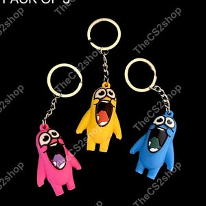 May include: A pack of 3 keychains featuring cartoon characters in pink, yellow, and blue. Each character has large white eyes, a wide open mouth, and is attached to a silver-toned metal split ring by a short chain. The text PACK OF 3 is visible.