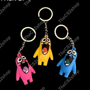 May include: Three keychains featuring cartoon characters in pink, yellow, and blue. Each character has wide eyes and an open mouth. The keychains include a metal ring and chain. The text PACK OF 1 is visible in the top left corner.