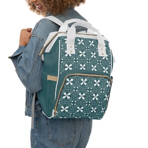 May include: A teal backpack featuring a white geometric pattern of four-petaled flowers and intersecting lines. The bag includes white top handles, a front zippered pocket, and gold-toned metal zippers. The side panels are solid teal with beige trim.