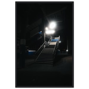Op de afbeelding: Een ingelijste verticale foto van een donkere trap in industriële stijl, verlicht door een fel licht. De treden hebben blauwe en witte accenten op de stootborden, omgeven door metalen leuningen tegen een diepzwarte achtergrond.