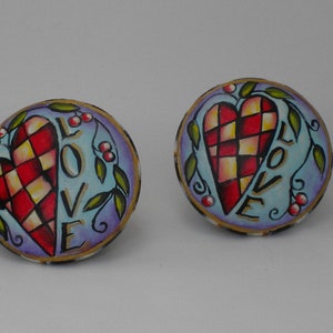May include: Four round, decorative, ceramic knobs with a painted design of a red and white checkered heart with the word "LOVE" written on it. The background is a light blue with a purple border and green vines with red berries.