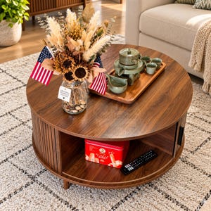 May include: A round walnut wood coffee table with a slatted base and an open storage shelf. On top sits a glass vase with dried flowers and two American flags, plus a wooden tray with a green ceramic tea set. A red tin and remote are below.