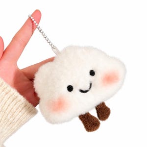 Op de afbeelding: Een witte pluche wolk sleutelhanger met een lachend gezicht met zwarte geborduurde ogen en mond, roze blosjes op de wangen en twee kleine bruine bungelende beentjes, bevestigd aan een zilverkleurige metalen ketting.