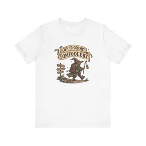 May include: A white t-shirt featuring a graphic of a small, cloaked figure with a backpack and lantern walking past a wooden signpost. Above the figure, a banner displays the text "OFF TO COMMIT TOMFOOLERY" in dark, stylised lettering.
