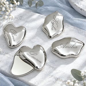 Peut inclure: Quatre miroirs de poche en métal argenté aux formes organiques irrégulières. Trois miroirs sont fermés, chacun gravé d'un prénom en écriture cursive : Olivia, Kylie et Amilia. Un miroir est ouvert, révélant une surface réfléchissante et un couvercle gravé du prénom Lauren.