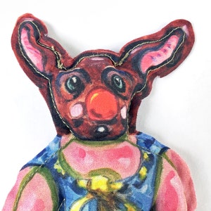 Puede incluir: Un juguete de peluche con una nariz roja y orejas grandes. El juguete lleva una camisa con estampado azul y amarillo.