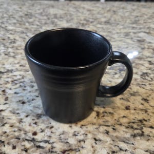 May include: A matte black ceramic mug with a slightly flared rim and a curved handle. The exterior surface features subtle horizontal ridges near the top, providing a textured appearance to the otherwise smooth, dark, solid-colored drinking vessel.