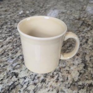 May include: A solid light cream-colored ceramic mug with a smooth, glossy finish. The mug features a cylindrical body, a rounded handle on the right side, and a subtle horizontal ring detail embossed near the top rim of the vessel.