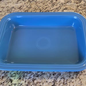 May include: A rectangular ceramic baking dish in a vibrant cornflower blue glaze. The dish features a smooth, glossy finish with a distinct tiered rim design and a small, concentric circular embossed detail centered on the interior base.