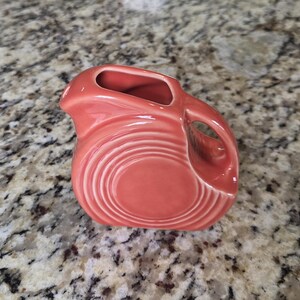 May include: A coral-colored ceramic creamer pitcher featuring a glossy finish and a series of concentric circular rings embossed on the side. The pitcher has a small pouring spout and a curved handle integrated into the side of the vessel.