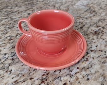 May include: A coral pink teacup and saucer set. The cup has a rounded shape and a handle. The saucer has a scalloped edge.
