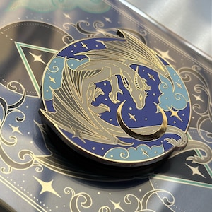 Può includere: Una spilla circolare in smalto raffigurante un drago grigio con le ali spiegate avvolto attorno a una mezzaluna dorata su uno sfondo blu intenso con nuvole azzurre e stelle dorate, posizionata su un cartoncino illustrato coordinato.