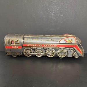 May include: A vintage tin toy train engine in dark gray with red and gold accents. The side features the text OVERLAND EXPRESS and the number 3140. A graphic of a Native American figure in a headdress is visible on the front section.