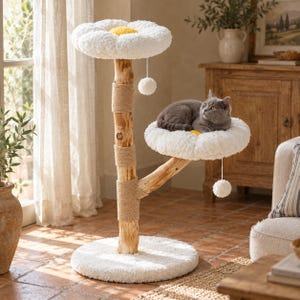 Puede incluir: Un árbol para gatos con un tronco de madera natural, secciones de cuerda de yute para rascar y dos plataformas blancas en forma de flor con centros amarillos. Un gato gris descansa en la plataforma inferior y dos juguetes de pompón cuelgan.