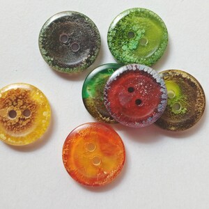 Puede incluir: Una colecci&oacute;n de seis botones circulares con dos orificios centrales cada uno. Los botones muestran variados patrones de colores transl&uacute;cidos y opacos, incluyendo tonos de verde oscuro, verde brillante, amarillo, naranja, rojo y marr&oacute;n texturizado.