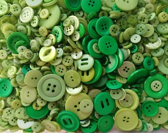 May include: A large collection of assorted sewing buttons in various shades of green, ranging from lime to forest green. The buttons vary in size, shape, and number of holes, with some featuring four holes, two holes, or slot-style openings.