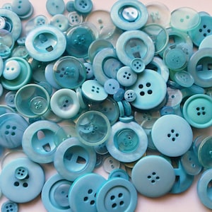 May include: A collection of various sizes of blue buttons. The buttons are in shades of turquoise, teal, and light blue. Some buttons have four holes, while others have two. The buttons are made of plastic and have a glossy finish.