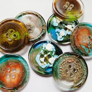 May include: A collection of seven round, four-hole buttons made of clear resin. Each button features a unique, swirling internal pattern of vibrant colors including orange, teal, green, brown, and white, creating a layered, depth-filled appearance.