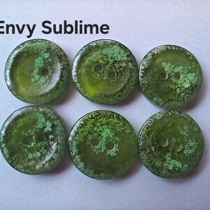 Puede incluir: Seis botones redondos de color verde con un dise&ntilde;o de dos agujeros y una superficie texturizada y moteada. El texto Envy Sublime est&aacute; impreso en negro sobre los botones. Cada bot&oacute;n tiene un acabado transl&uacute;cido con tonos verde bosque.