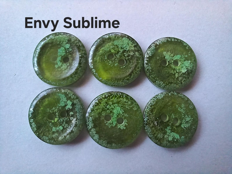 Puede incluir: Seis botones redondos de color verde con un dise&ntilde;o de dos agujeros y una superficie texturizada y moteada. El texto Envy Sublime est&aacute; impreso en negro sobre los botones. Cada bot&oacute;n tiene un acabado transl&uacute;cido con tonos verde bosque.