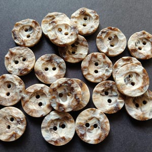 May include: A collection of round buttons with two holes each. The buttons have a marbled appearance with shades of brown and cream, and a slightly textured surface. These buttons are suitable for sewing and craft projects.