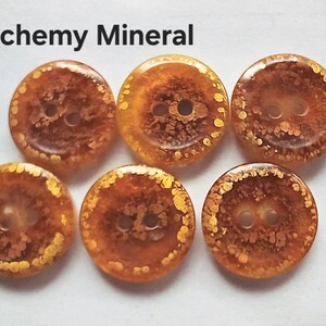 Puede incluir: Cinco botones redondos con un patr&oacute;n transl&uacute;cido de color &aacute;mbar y motas doradas, cada uno con dos agujeros centrales. El texto Alchemy Mineral est&aacute; impreso en negro sobre los botones. Los botones tienen un acabado liso y brillante de tonos c&aacute;lidos.