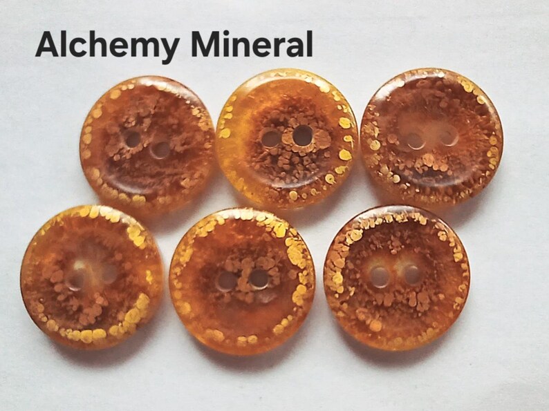 Puede incluir: Cinco botones redondos con un patr&oacute;n transl&uacute;cido de color &aacute;mbar y motas doradas, cada uno con dos agujeros centrales. El texto Alchemy Mineral est&aacute; impreso en negro sobre los botones. Los botones tienen un acabado liso y brillante de tonos c&aacute;lidos.