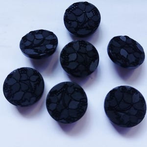 May include: A set of seven round black buttons, each featuring a raised floral pattern on the surface. The buttons have a smooth, dark finish with intricate petal and leaf details embossed across the top of every individual circular piece.