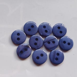May include: A collection of ten round, two-hole buttons in a solid periwinkle blue color. Each button features a smooth, slightly glossy finish with a subtle marbled texture throughout the surface, arranged in a loose cluster on a plain background.