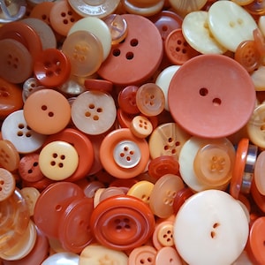 May include: A collection of various orange, peach, and cream-colored buttons. The buttons vary in size and shape, with two-hole and four-hole designs. Some buttons have a glossy finish, while others are matte.