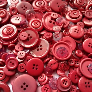 May include: A collection of various red buttons of different sizes and shapes. The buttons have two or four holes and are made of plastic. Some buttons are solid red, while others have a slightly translucent appearance.