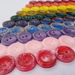May include: A rainbow assortment of colorful buttons with a glittery finish. The buttons are arranged in a diagonal line, starting with red and ending with purple.