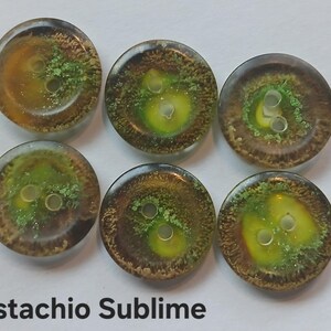 Puede incluir: Seis botones redondos con un acabado liso y brillante, que presentan una mezcla arremolinada de pigmentos &aacute;mbar transl&uacute;cidos, marr&oacute;n oscuro y verde vibrante. Cada bot&oacute;n tiene dos agujeros centrales y el texto Pistachio Sublime est&aacute; impreso debajo.