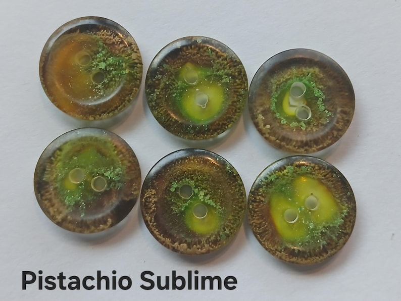 Puede incluir: Seis botones redondos con un acabado liso y brillante, que presentan una mezcla arremolinada de pigmentos &aacute;mbar transl&uacute;cidos, marr&oacute;n oscuro y verde vibrante. Cada bot&oacute;n tiene dos agujeros centrales y el texto Pistachio Sublime est&aacute; impreso debajo.