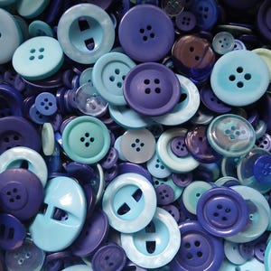 May include: A large collection of assorted sewing buttons in various shades of blue, ranging from light sky blue to deep navy. The buttons feature different designs, including standard four-hole styles and two-hole toggle shapes in various sizes.
