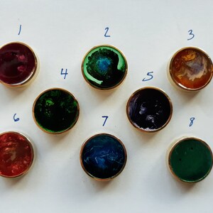 May include: Eight round, gold-rimmed decorative knobs with colorful, swirled resin centers. The colors include shades of red, green, blue, and purple. The knobs are arranged on a white surface.