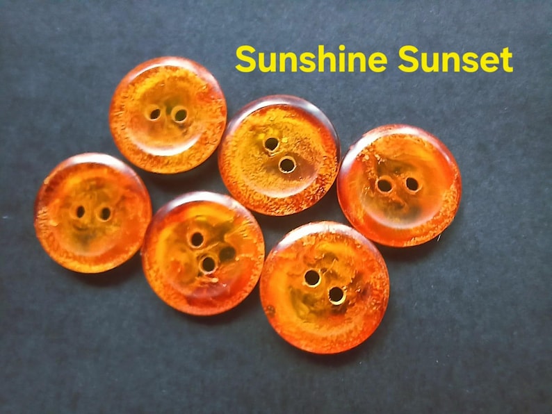 Puede incluir: Seis botones redondos con un patr&oacute;n jaspeado de color &aacute;mbar y naranja transl&uacute;cido, cada uno con dos agujeros centrales. El texto Sunshine Sunset est&aacute; impreso en amarillo sobre los botones. Los botones tienen un acabado liso y brillante.