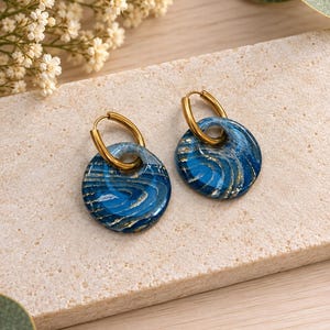 May include: A pair of hoop earrings featuring circular blue resin charms with swirling white and gold-toned metallic accents. The charms are suspended from polished gold-toned metal hoops, creating a cohesive set of jewelry with a glossy finish.