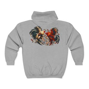 Peut inclure: Un sweat à capuche zippé gris clair présentant un motif détaillé au dos représentant deux coqs dans une posture dynamique. Les coqs sont rendus dans des tons vifs de rouge, orange, noir et crème, entourés de plumes et de débris.
