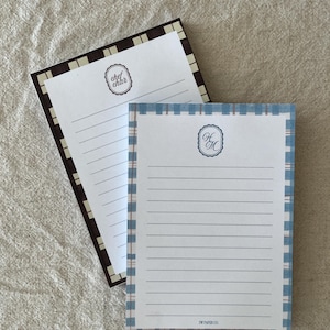 May include: Two rectangular lined notepads with plaid borders. The top notepad has a blue plaid border and the text HM inside an oval frame. The bottom notepad has a dark brown plaid border and the text chef char inside an oval frame.
